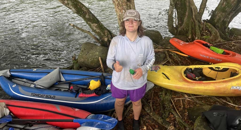 A person standing on the bank of a river near several beached watercraft gives a thumbs up